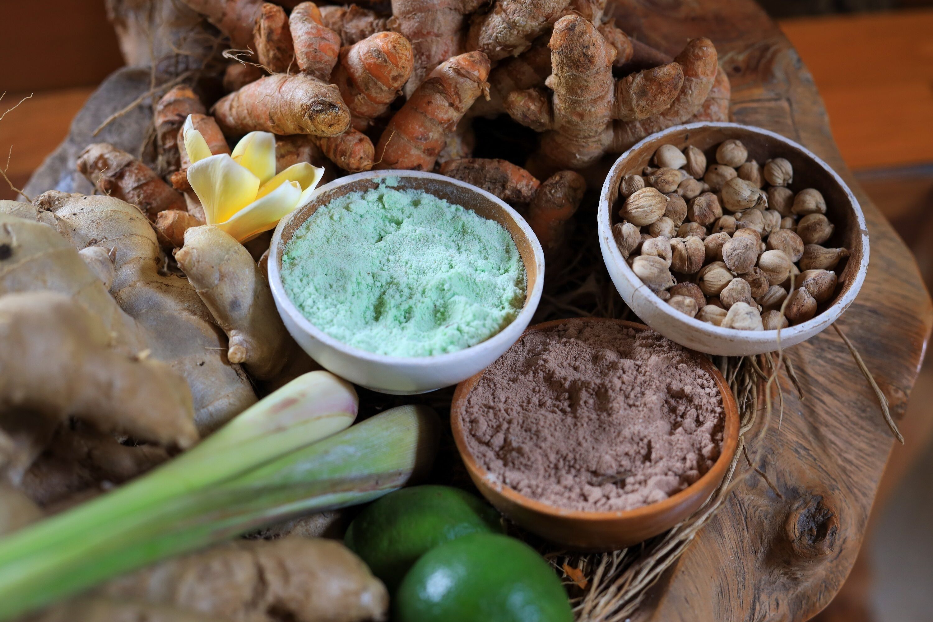 A wooden table with ginger roots, green leaves, and two bowls of spices.