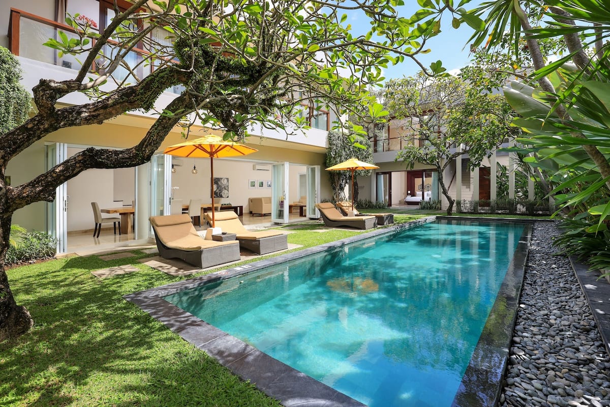 A swimming pool with lounge chairs and umbrellas, surrounded by greenery and a modern building.