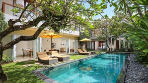 Una piscina con sillones de descanso y sombrillas, rodeada de vegetación y un edificio moderno.
