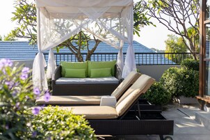 A covered outdoor seating area with a wicker sofa and cushions, surrounded by greenery and a tree.