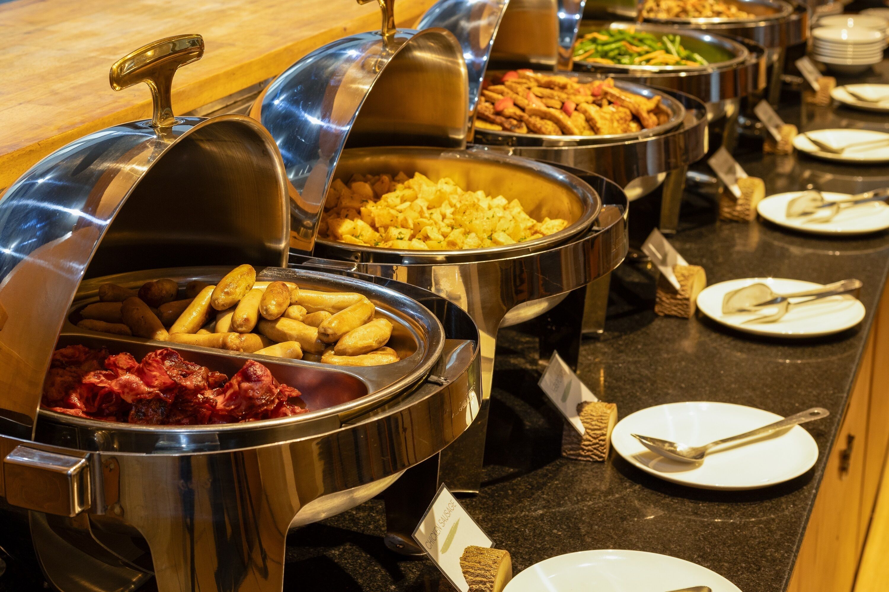A buffet with various food items in metal containers, including sausages, potatoes, and vegetables.