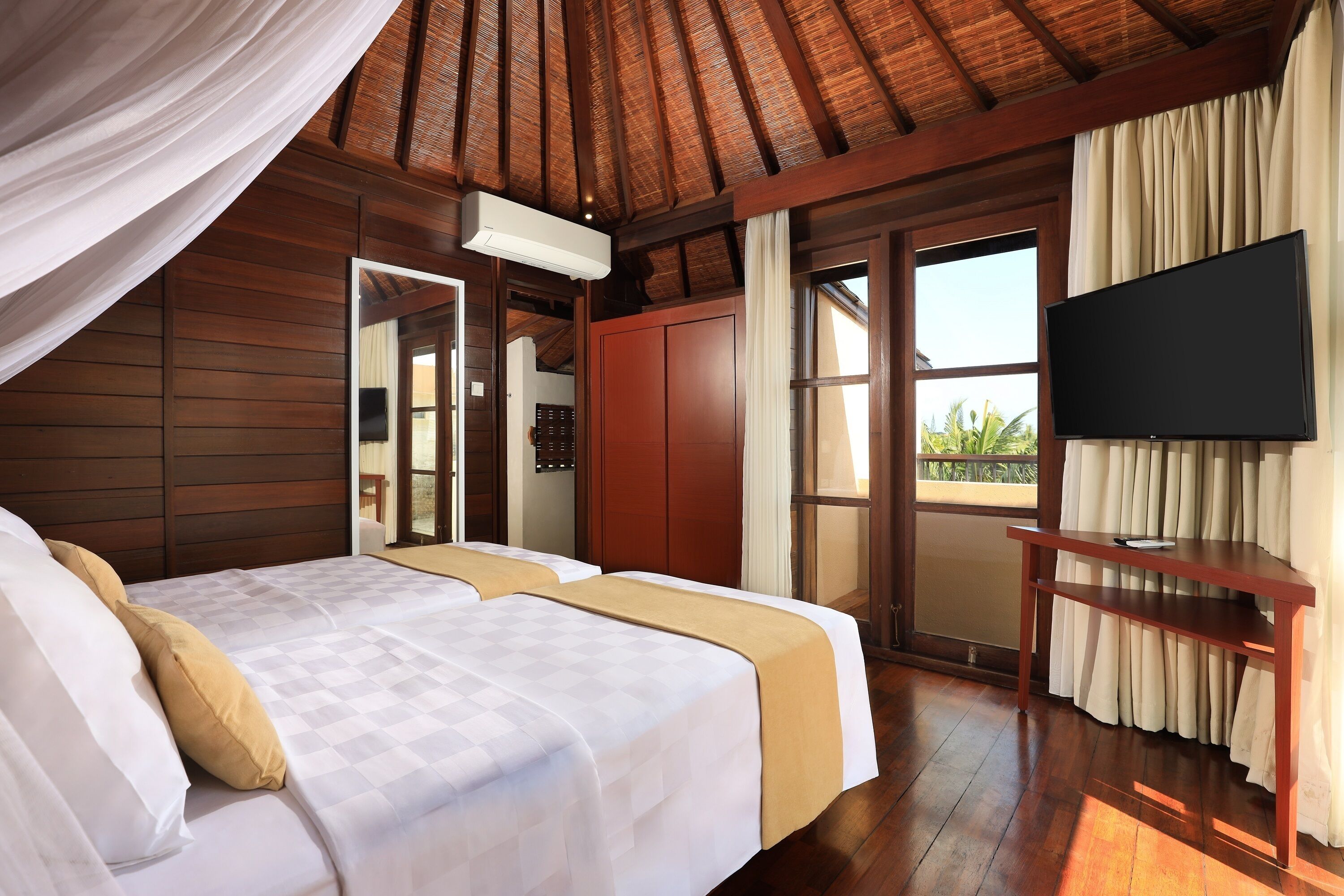 Habitación de hotel con techo de madera, cama con ropa de cama blanca y color beige, televisor sobre un mueble de madera y vista al exterior a través de un ventanal grande.