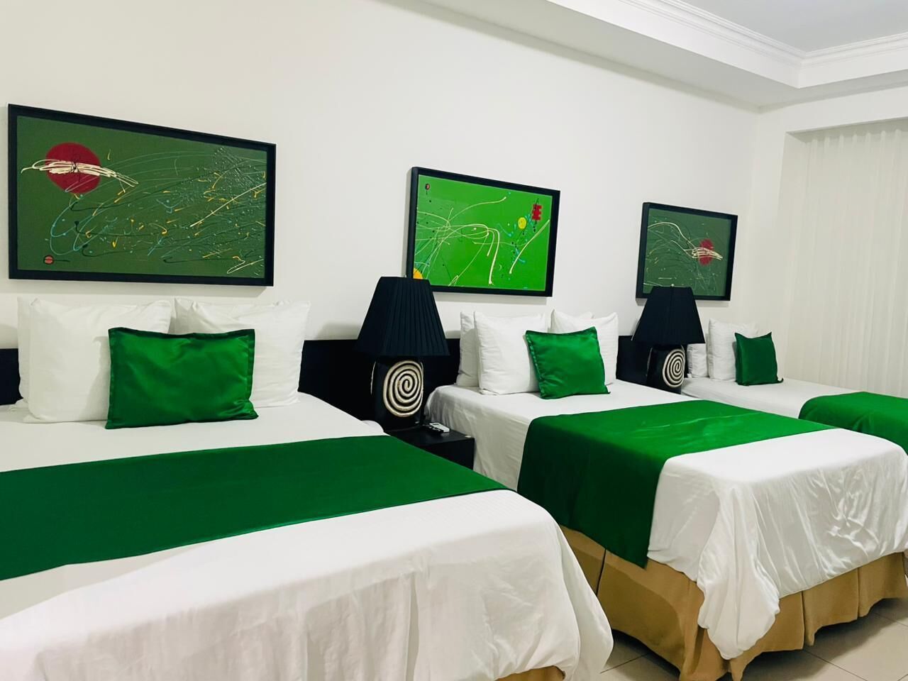 Quarto de hotel com três camas, cada uma com colcha verde e roupa de cama branca. Há três quadros abstratos nas paredes, e uma luminária preta com design em espiral no criado-mudo.
