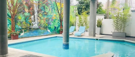 An indoor swimming pool with a mural, lounge chairs, and potted plants.