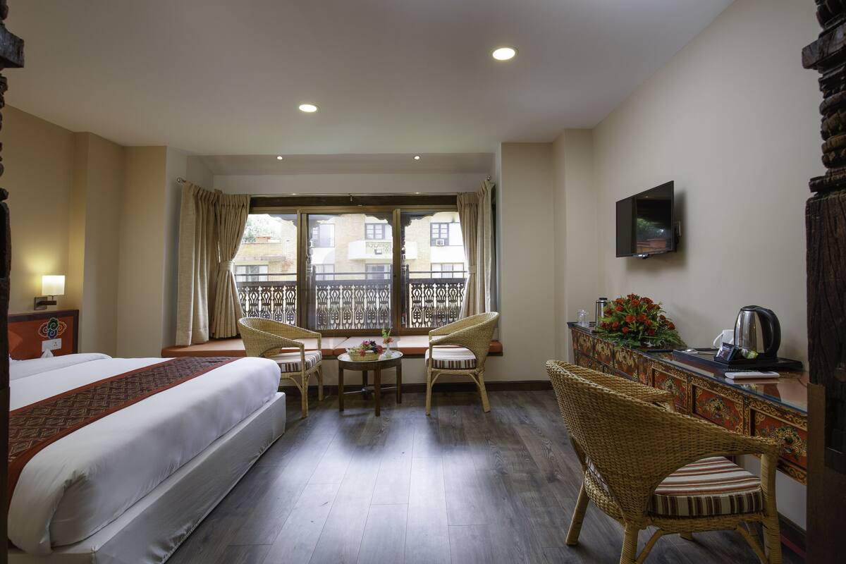 Habitación de hotel con cama, una pequeña mesa con sillas, un balcón con vistas a edificios y un televisor montado en la pared.