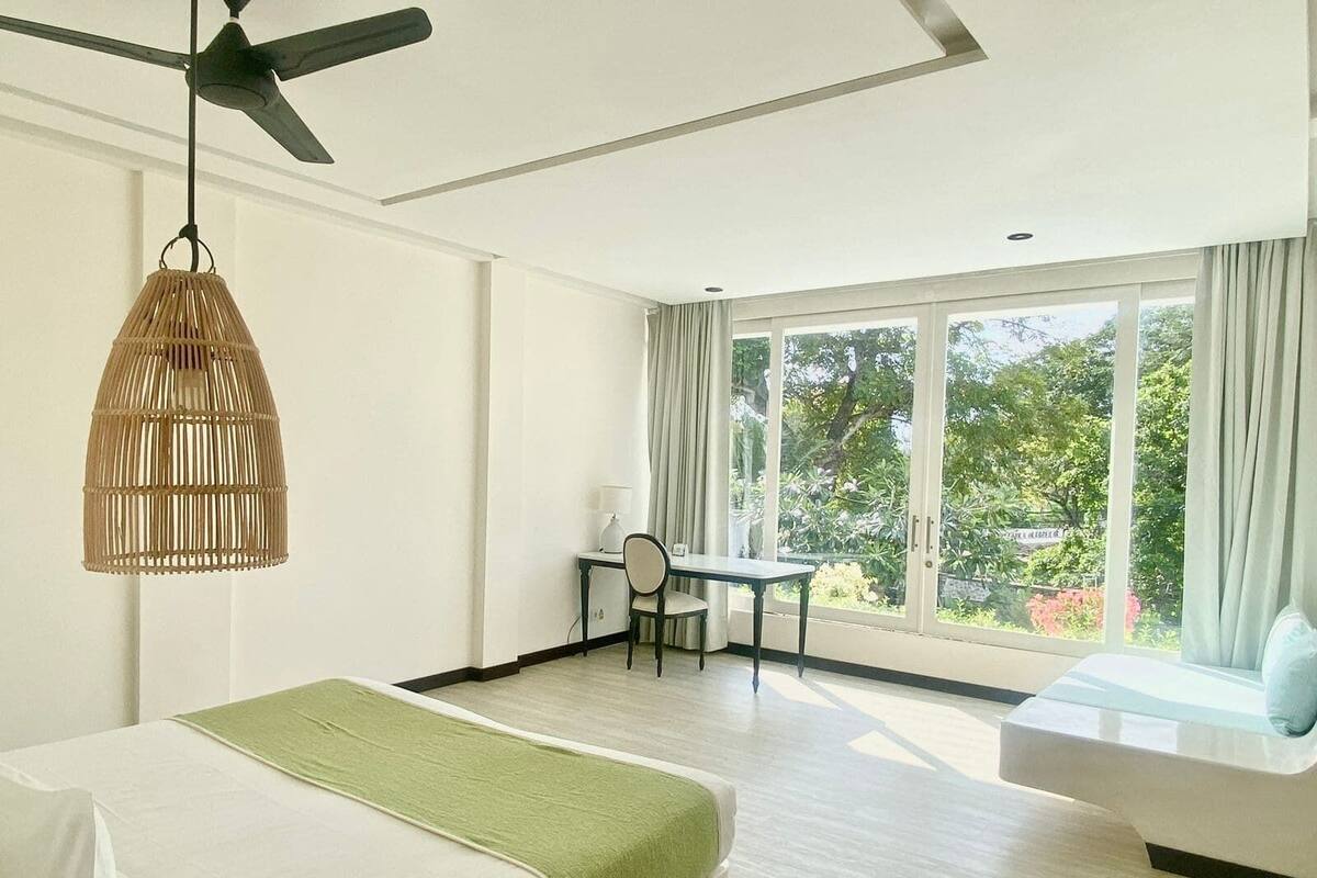 Una camera d'albergo con un letto, una scrivania, una sedia, un divano, una ventola a soffitto e ampie finestre.