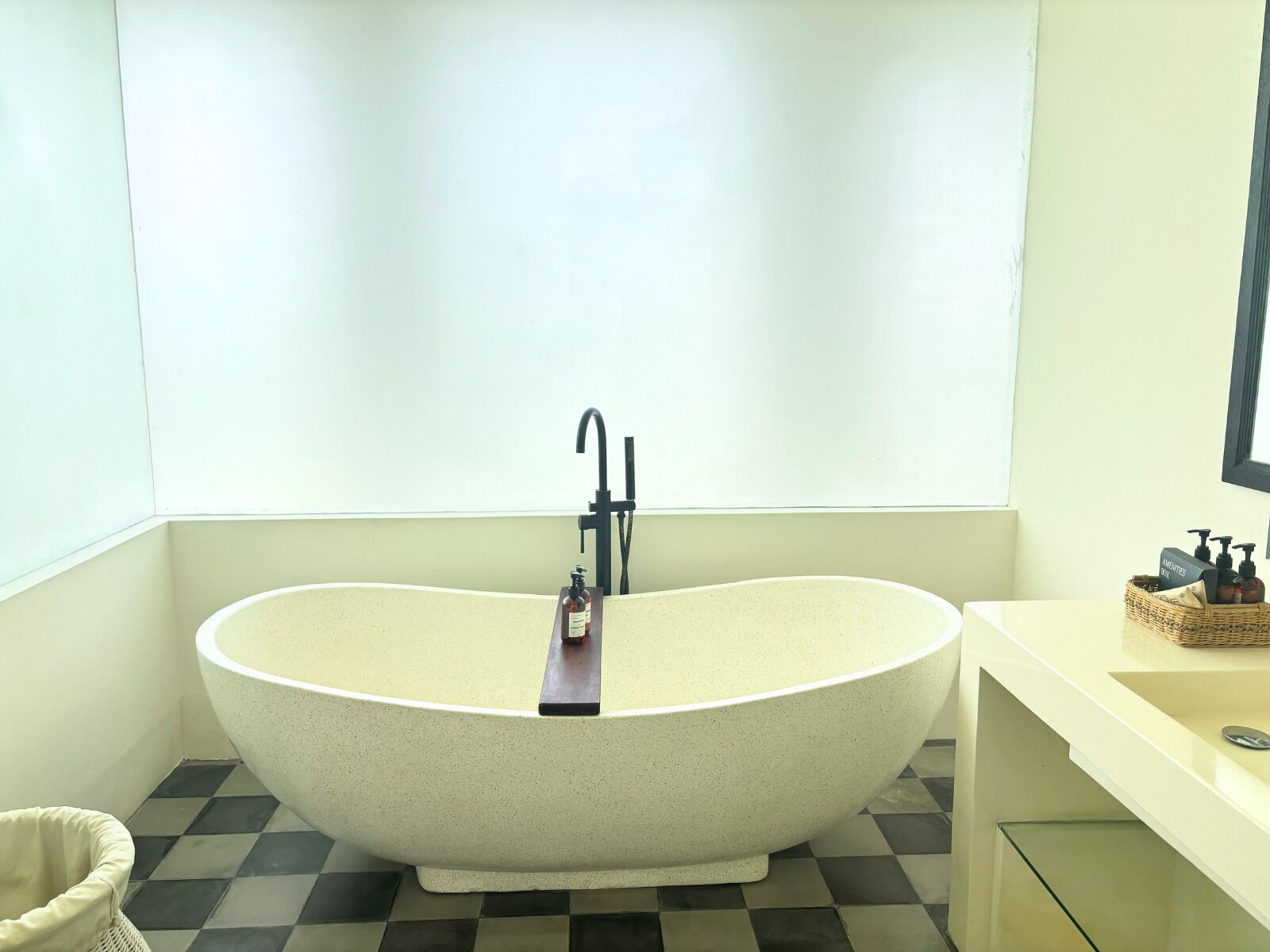 A freestanding bathtub in a bathroom with a checkered floor, a small wicker basket with toiletries, and a mirror on the wall.