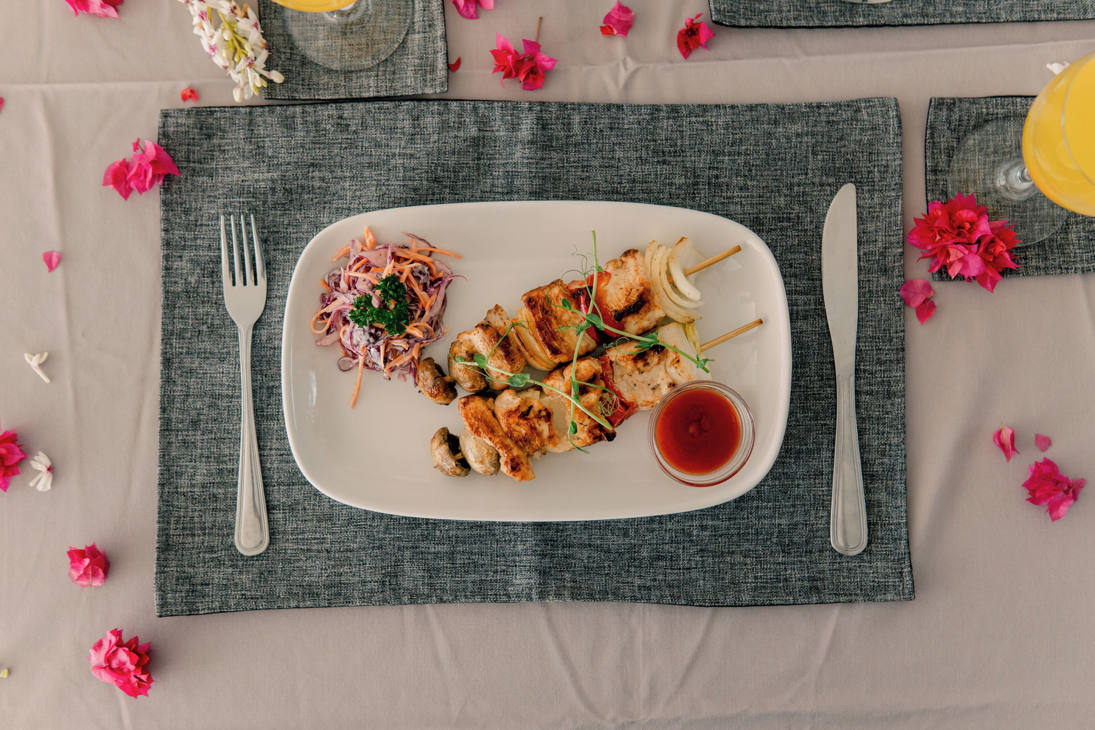 A neatly arranged meal on a white plate with a side salad, skewers, and a small bowl of sauce, set on a table with a gray placemat and scattered pink flowers.