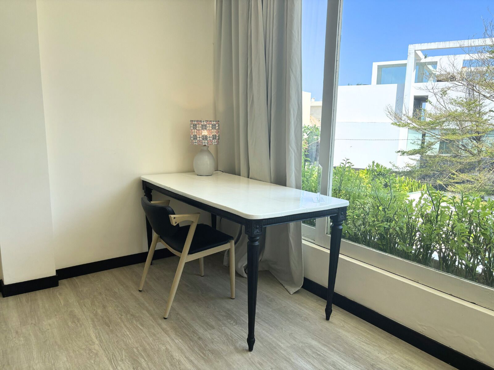 Una habitación con una mesa blanca, una silla negra, una lámpara con diseño y vistas a zonas verdes y edificios.