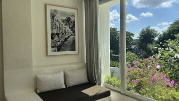 A room with a large window, a black and white framed picture on the wall, a white sofa with cushions, and a view of greenery and flowers outside.
