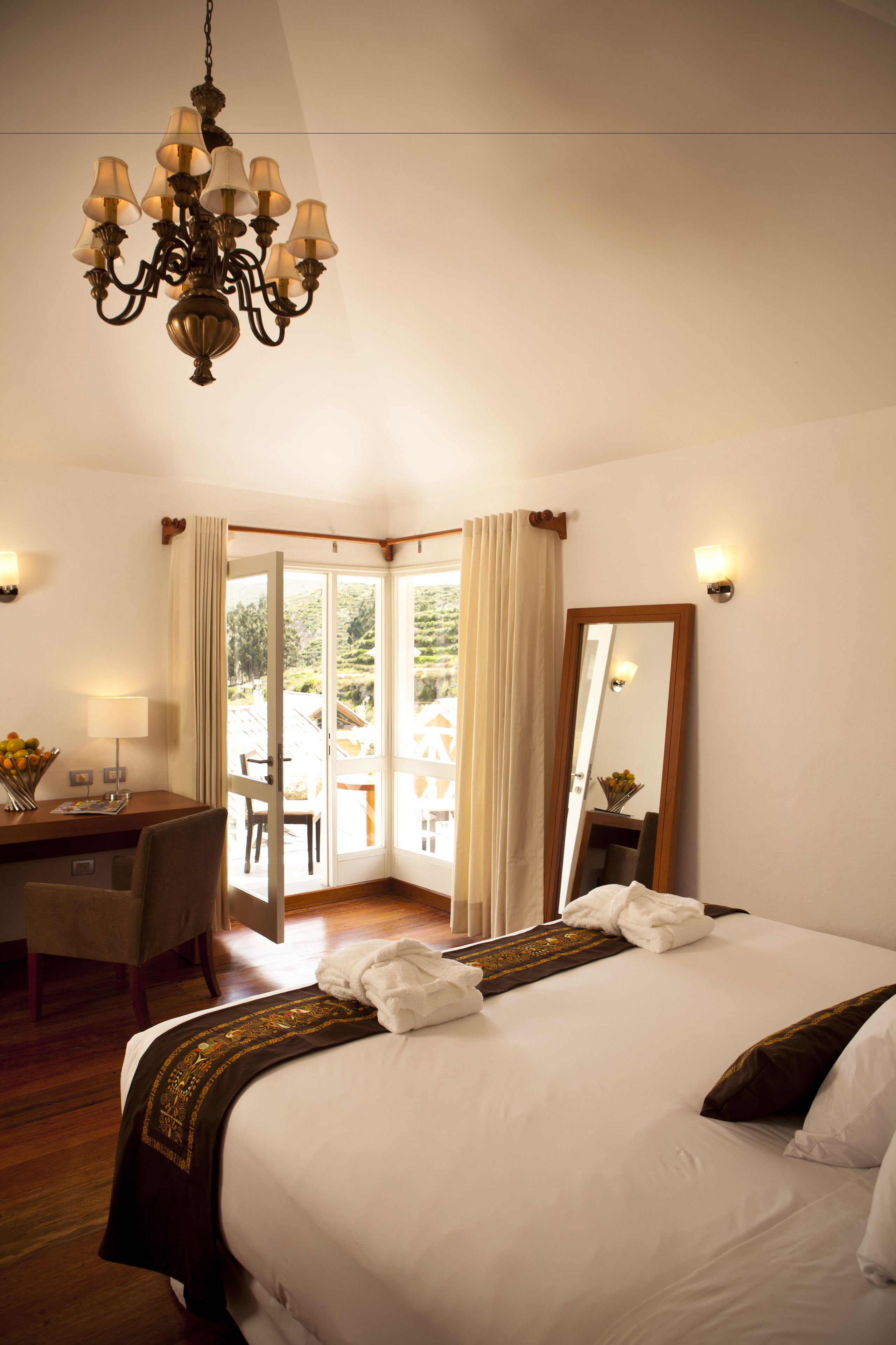 A hotel room with a large bed, a desk with a chair, a chandelier, and a view through a glass door.