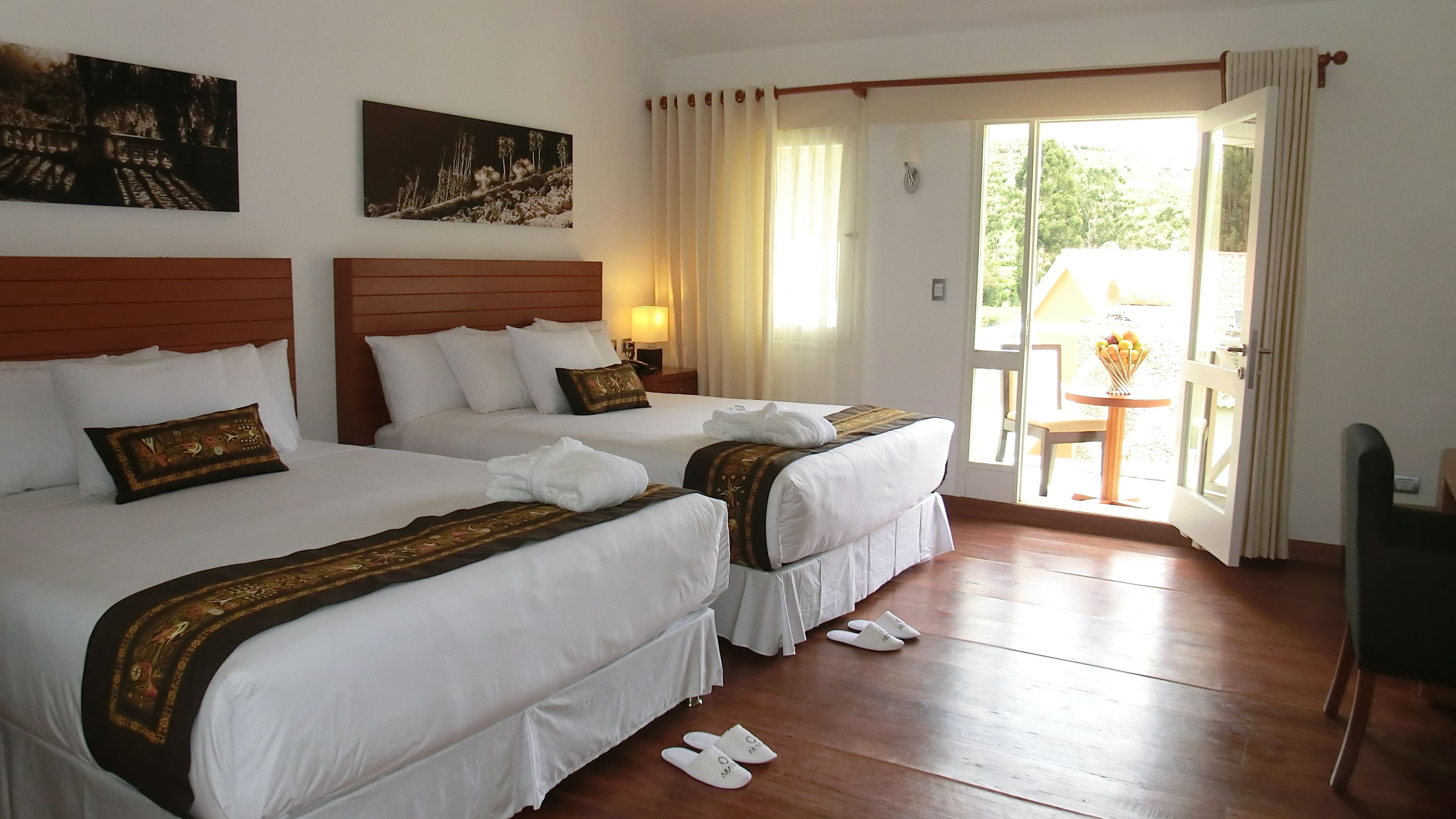 Une chambre d’hôtel avec deux lits, une tête de lit en bois, du linge de lit blanc, une petite table avec une lampe et une vue sur l’extérieur par une porte.