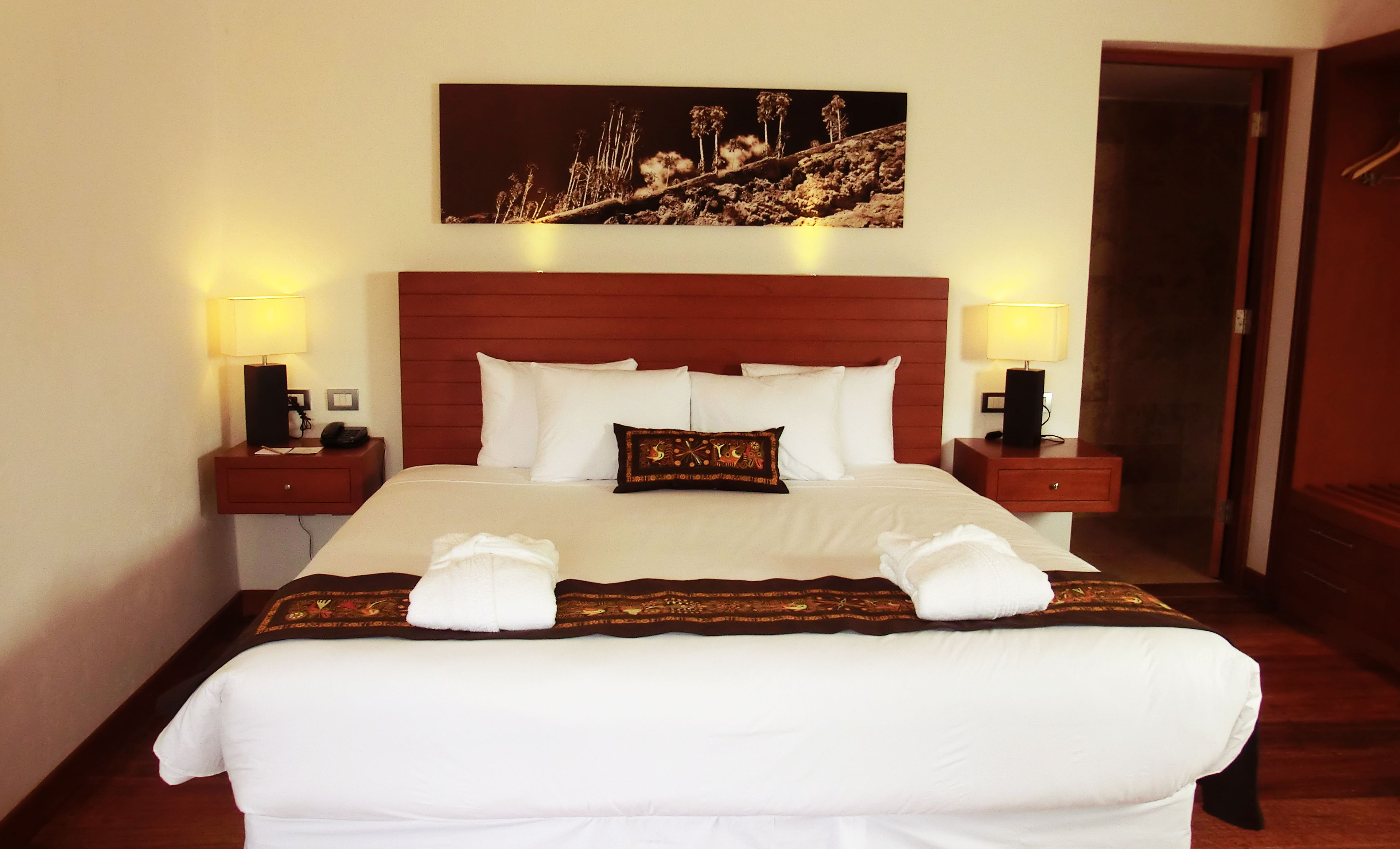 A neatly made bed with white linens and a decorative pillow, flanked by two bedside tables with lamps, in a room with a wooden headboard and a framed picture above the bed.