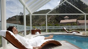 Une personne se détend dans un fauteuil en bois au bord d’une piscine, un verre de jus d’orange à la main, et est entourée d’une structure en verre offrant une vue sur les montagnes.