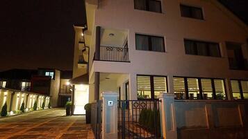 Un edificio de varios pisos con entrada protegida por una reja, un balcón en el segundo nivel y una fachada bien iluminada.