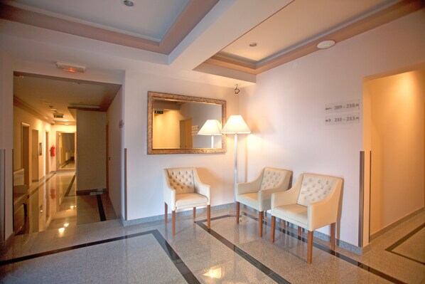 A hotel lobby with a polished marble floor, two upholstered chairs, a wall-mounted mirror, and a floor lamp.