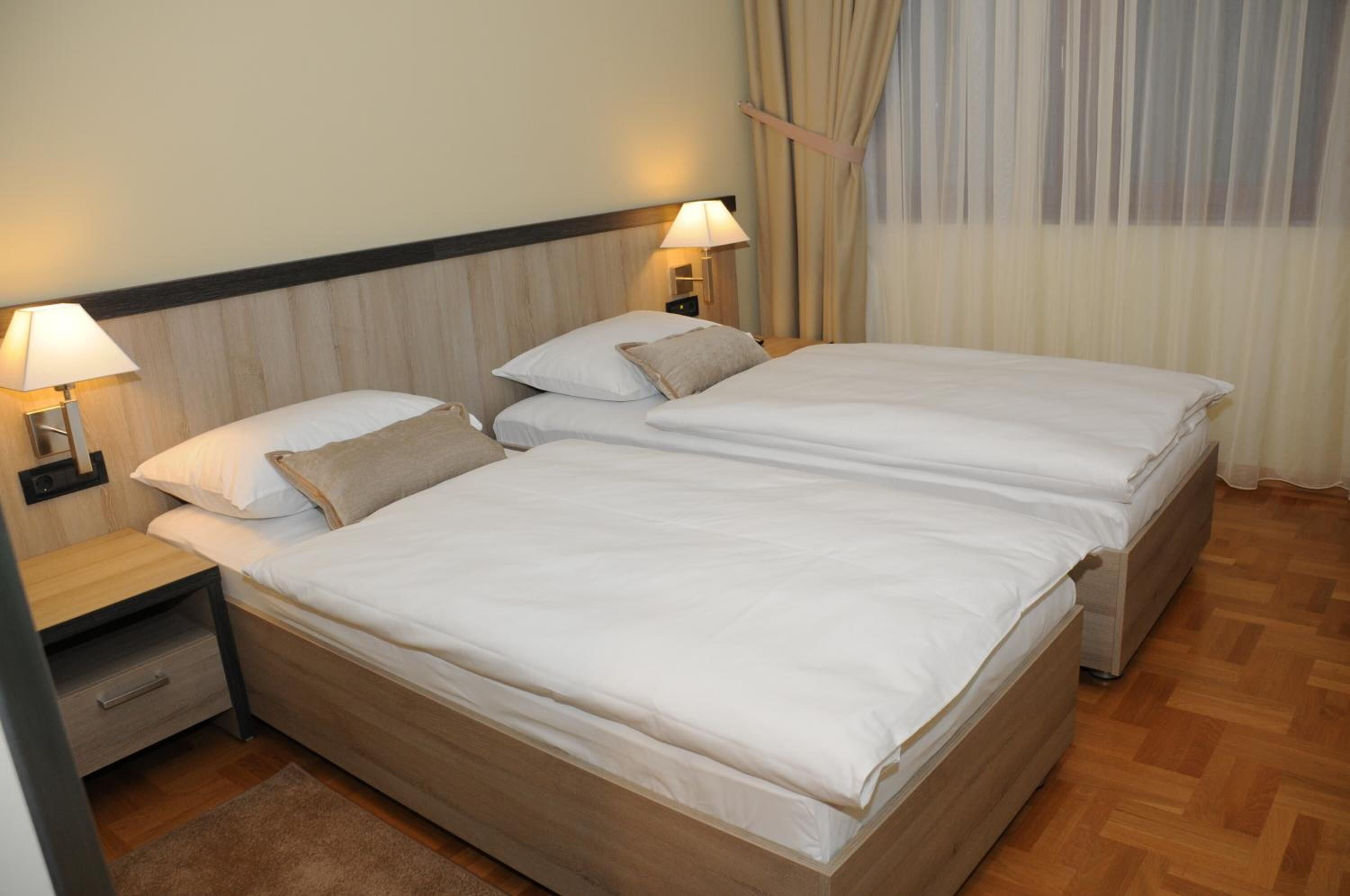A double bed with two bedside tables, each with a lamp, in a room with wooden flooring and a window with curtains.