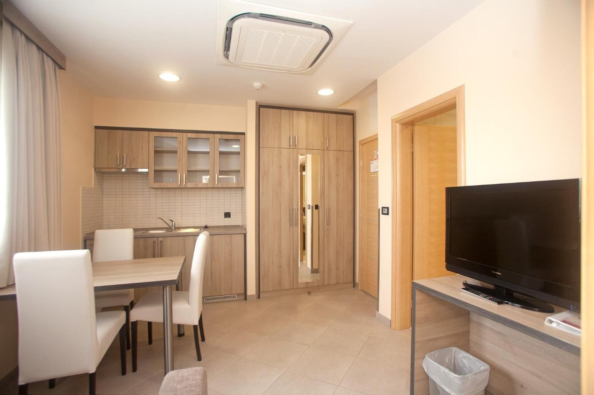A compact kitchen with wooden cabinets, a dining area with white chairs, a television on a stand, and a trash bin.