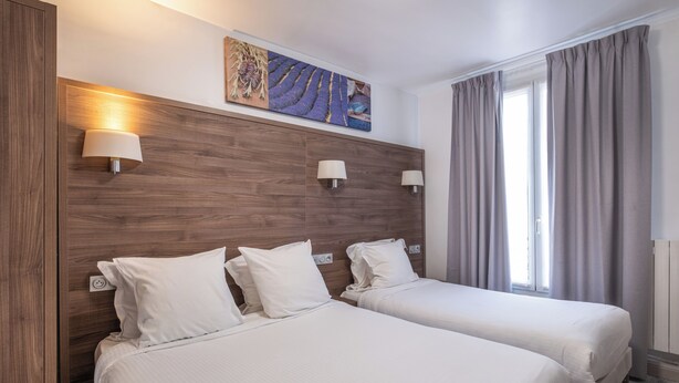 A hotel room with two beds, a wooden headboard, wall-mounted lamps, a painting, and a window with curtains. - Hôtel Luxor (Issy-les-Moulineaux)