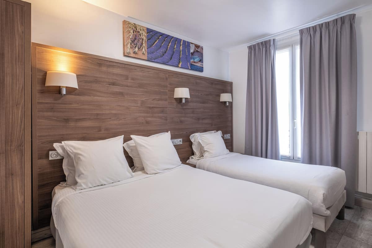 A hotel room with two beds, a wooden headboard, wall-mounted lamps, a painting, and a window with curtains.