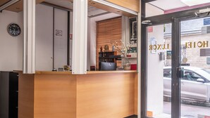 A hotel reception area with a wooden counter, a clock, and a view of the street outside. - Hôtel Luxor (Issy-les-Moulineaux)