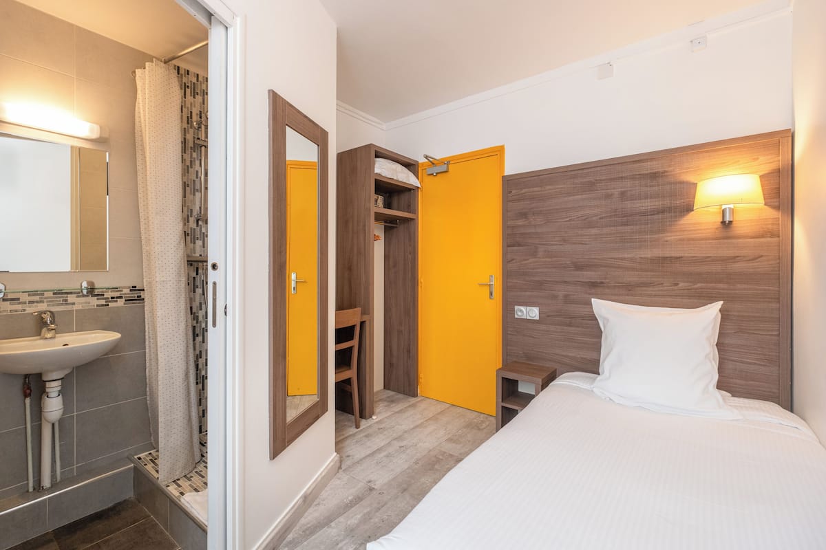 A hotel room with a wooden headboard, a white bed, a wooden wardrobe, a bathroom with a sink and mirror, and a shower area.