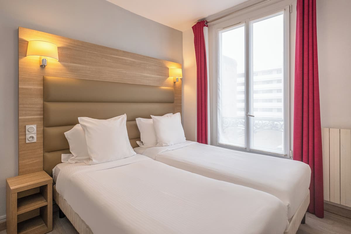 A hotel room with two beds, a wooden headboard, bedside lamps, a nightstand, and a window with red curtains.