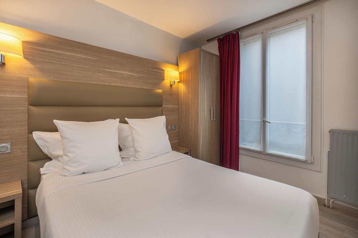 A hotel room with a large bed, a wooden headboard, a bedside table, a lamp, and a window with curtains.