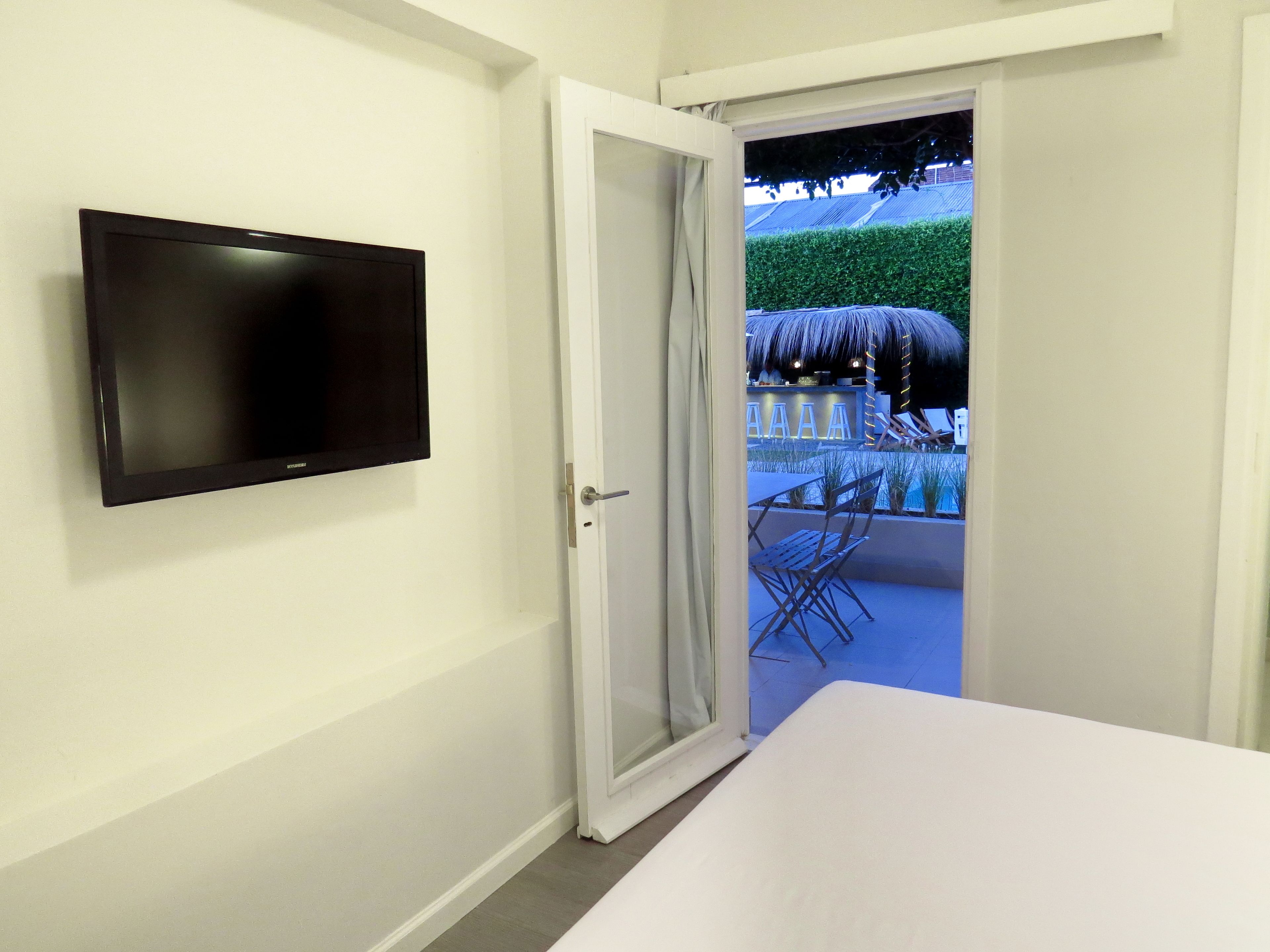 A room with a TV mounted on the wall, a bed, and an open door leading to an outdoor area with a pool and lounge chairs.