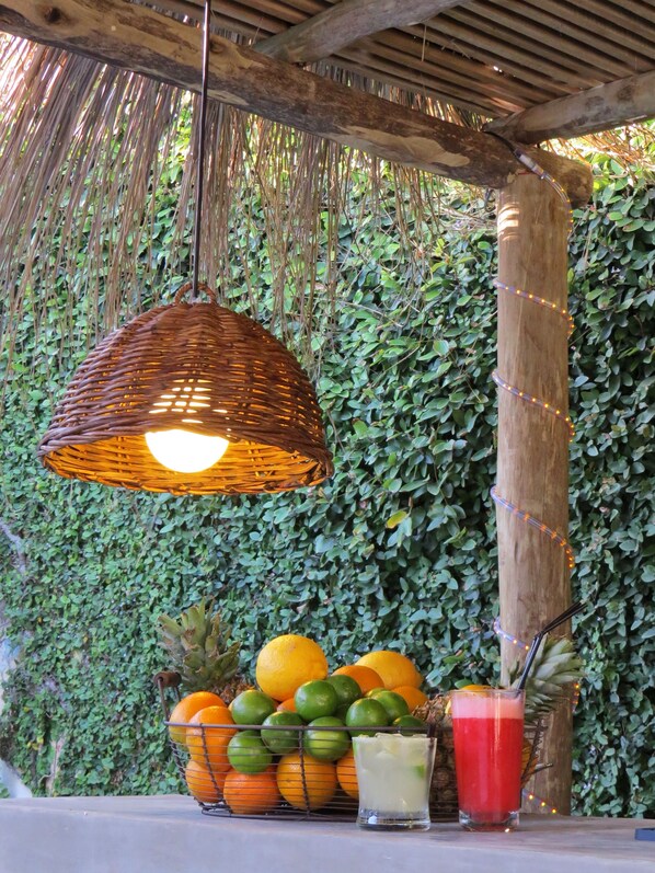 Una lámpara colgante de mimbre, una cesta de frutas con naranjas y limones, y dos vasos de bebidas sobre una mesa.