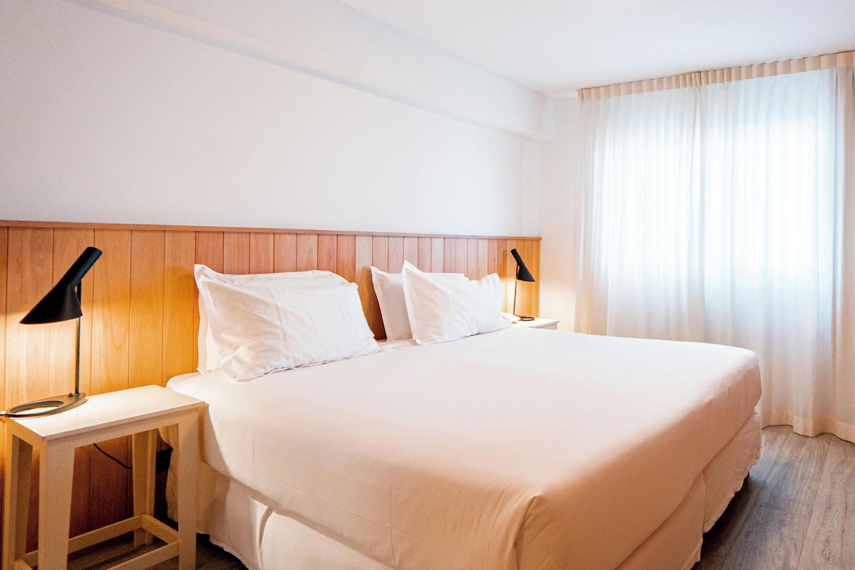 Une chambre d’hôtel avec un grand lit, deux tables de chevet avec des lampes, un téléviseur fixé au mur et une fenêtre avec des rideaux.