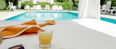 Una mesa con un plato de comida, una bebida y gafas de sol, con vista a una piscina con sillas de descanso y follaje.