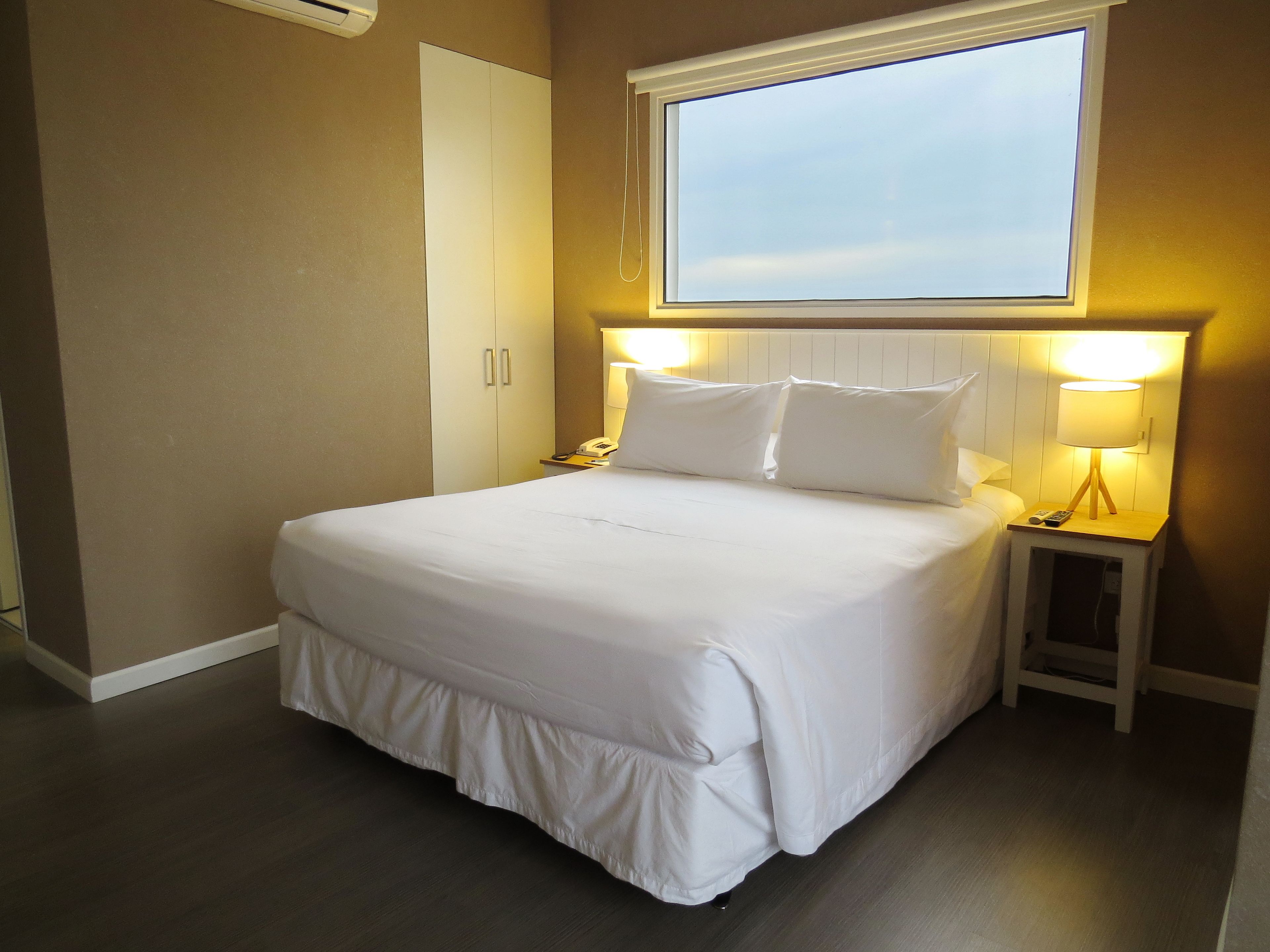 A neatly made bed with white bedding, a bedside table with a lamp, and a window above the bed.