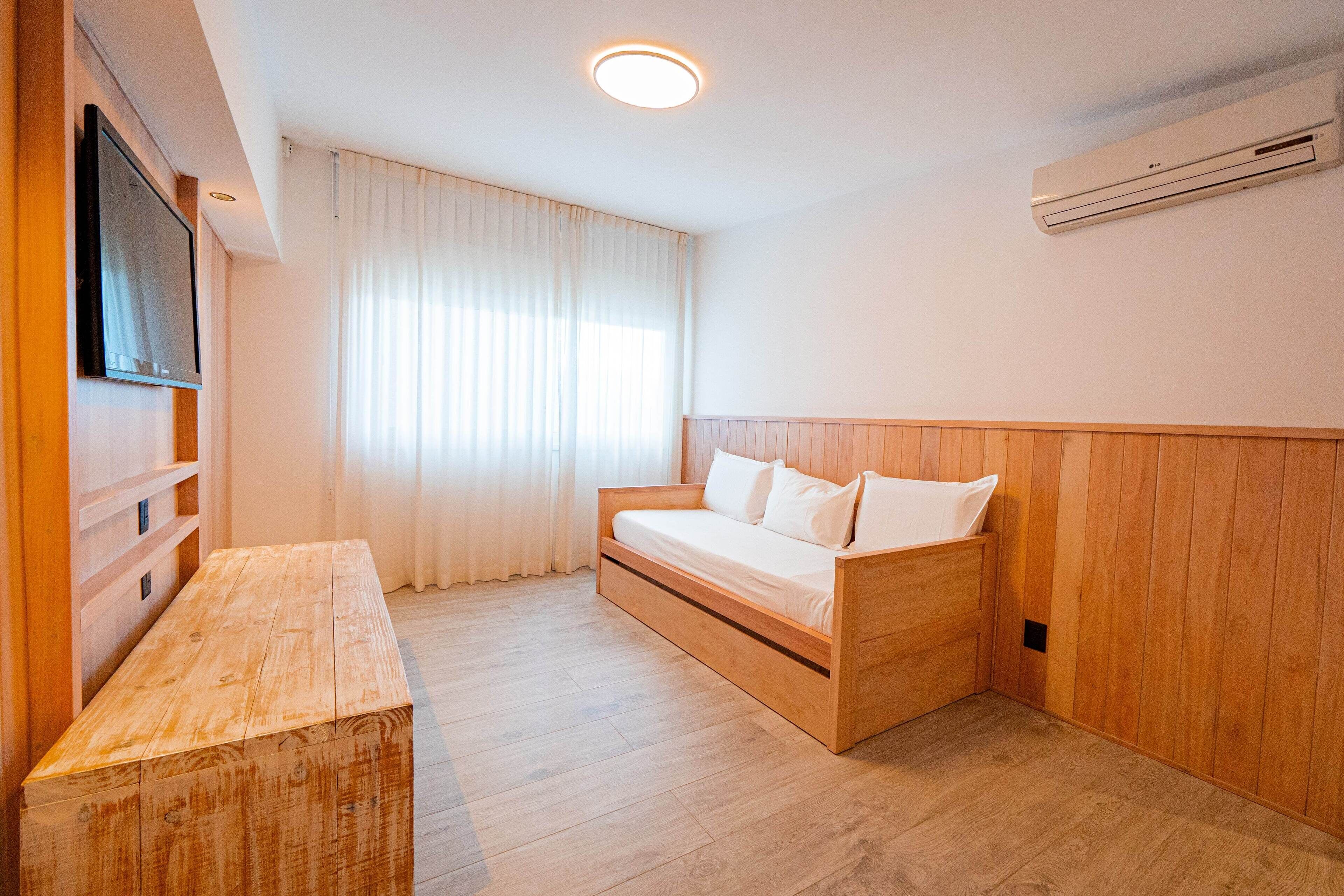 Ein Zimmer mit einem hölzernen Bett, einer Holzbank, einem Flachbildfernseher und einem Fenster mit Vorhängen.