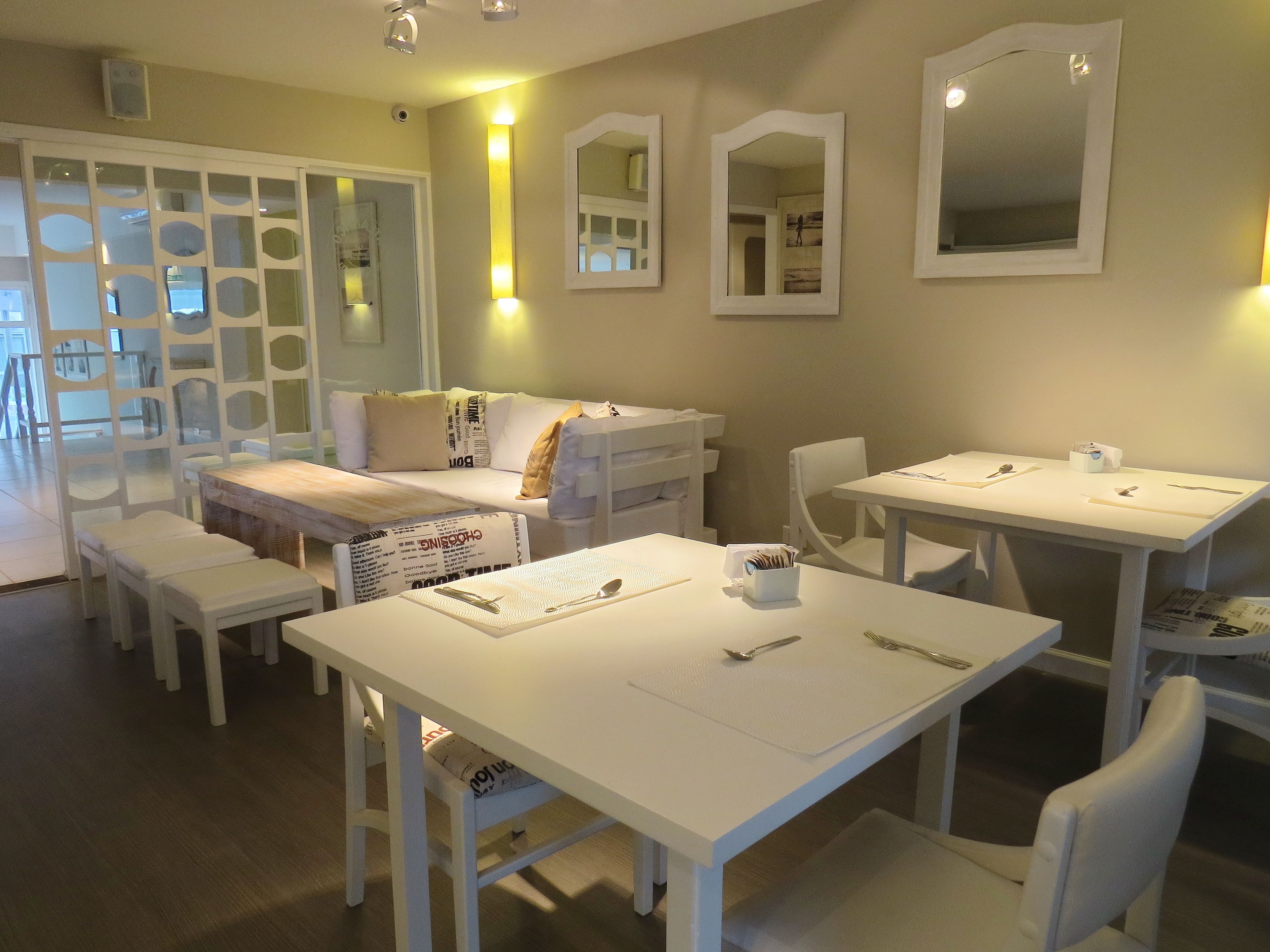 A modern, well-lit room with a dining table set for two, a sofa, and a white-framed mirror on the wall.