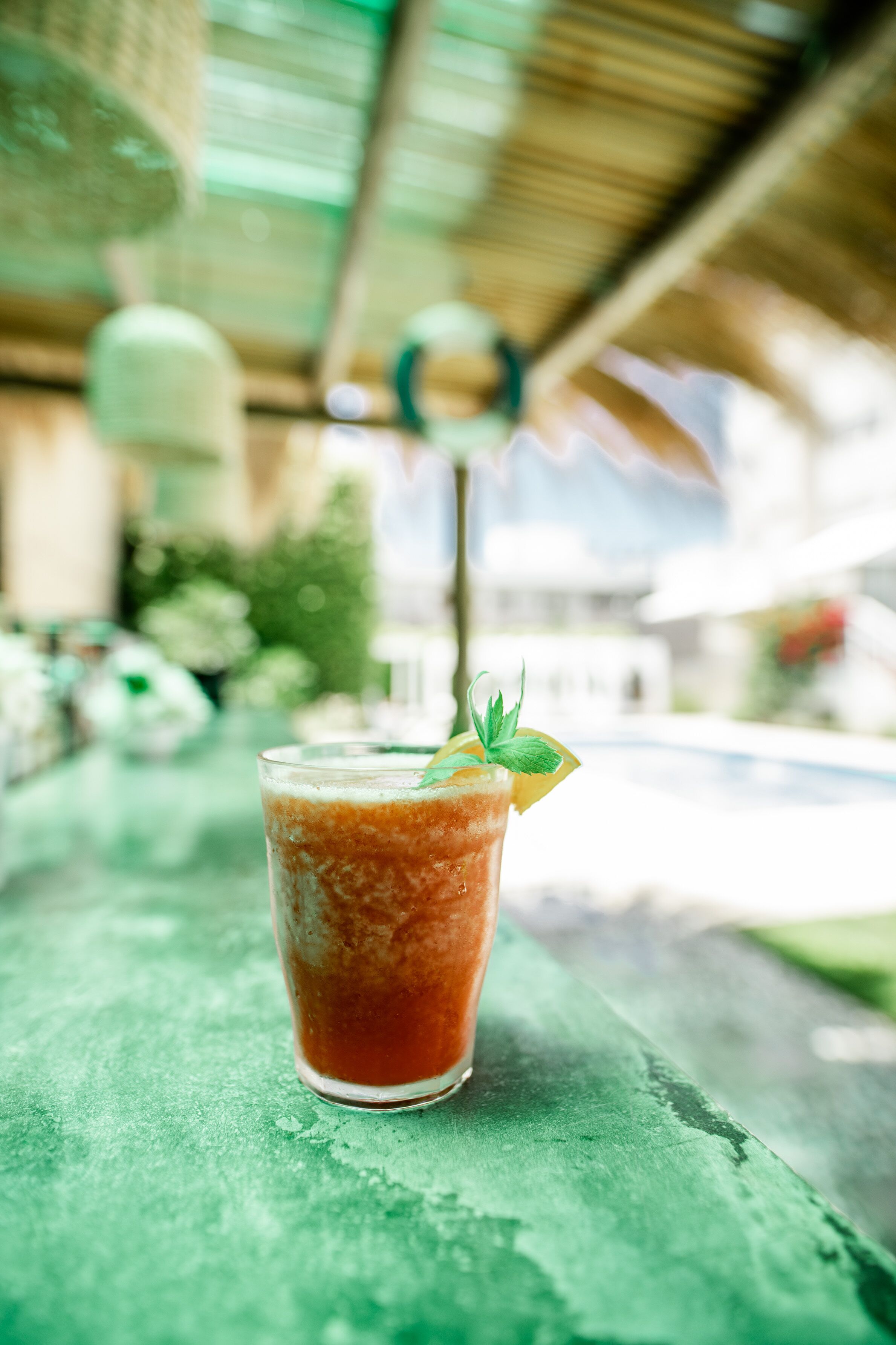 A glass of iced beverage with a mint garnish on a green table under a thatched roof.