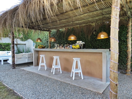 Zona de bar al aire libre con techo de paja, tres taburetes blancos y luces colgantes.