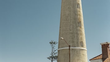 Un alto faro cilíndrico con la parte superior roja y la base blanca, rodeado de palmeras y edificios.