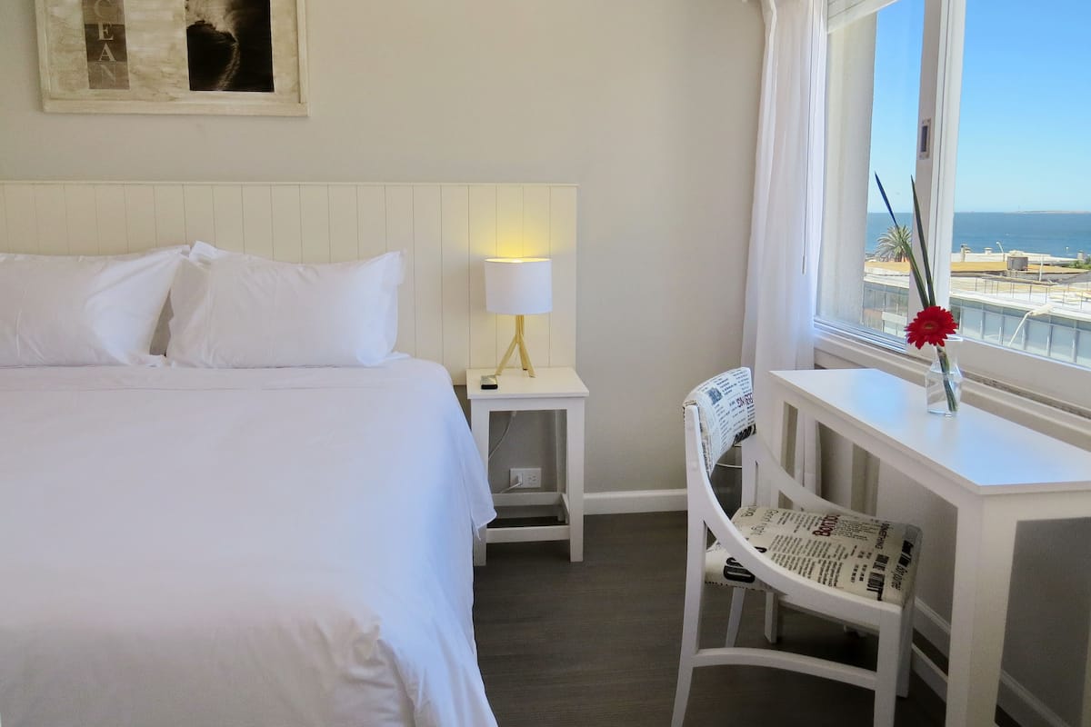 Une chambre d’hôtel avec un lit, un bureau doté d’une lampe, une chaise, une fenêtre donnant sur la mer et un tableau encadré au mur.