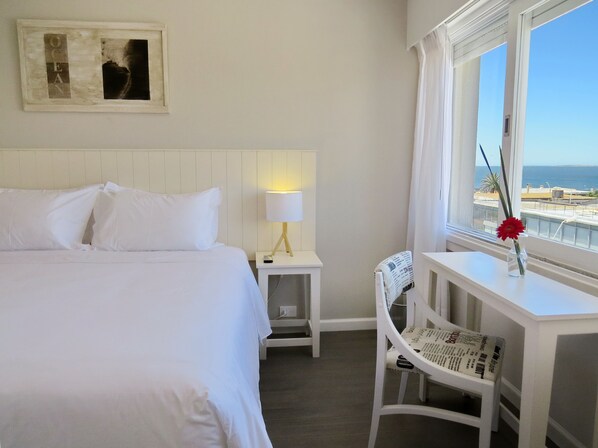 A hotel room with a bed, a desk with a lamp, a chair, a window with a view of the sea, and a framed picture on the wall. - Atlántico Boutique Hotel (Punta del Este)