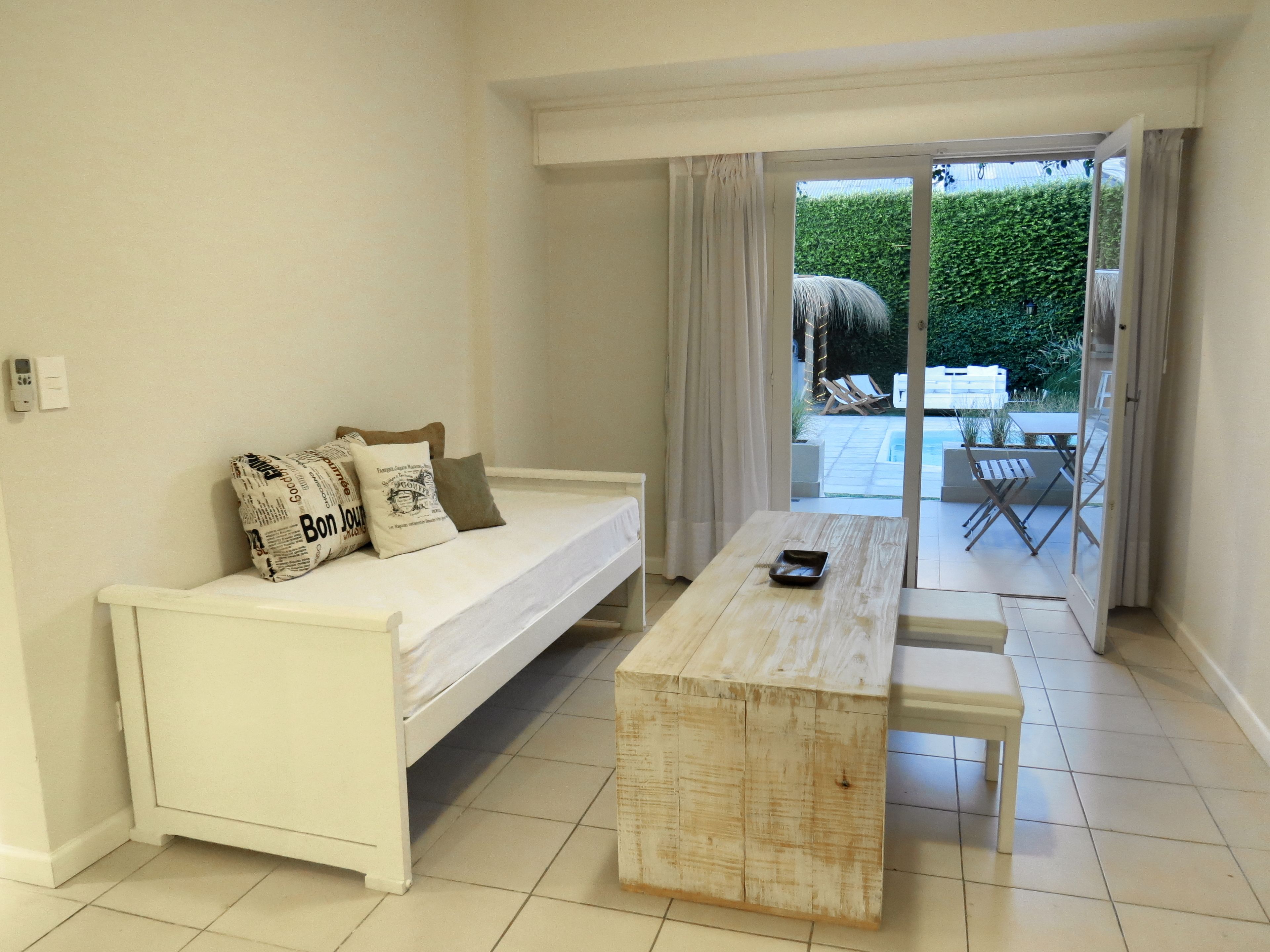 A minimalist room with a white sofa, a wooden table, and a sliding glass door leading to an outdoor area with a pool and lounge chairs.