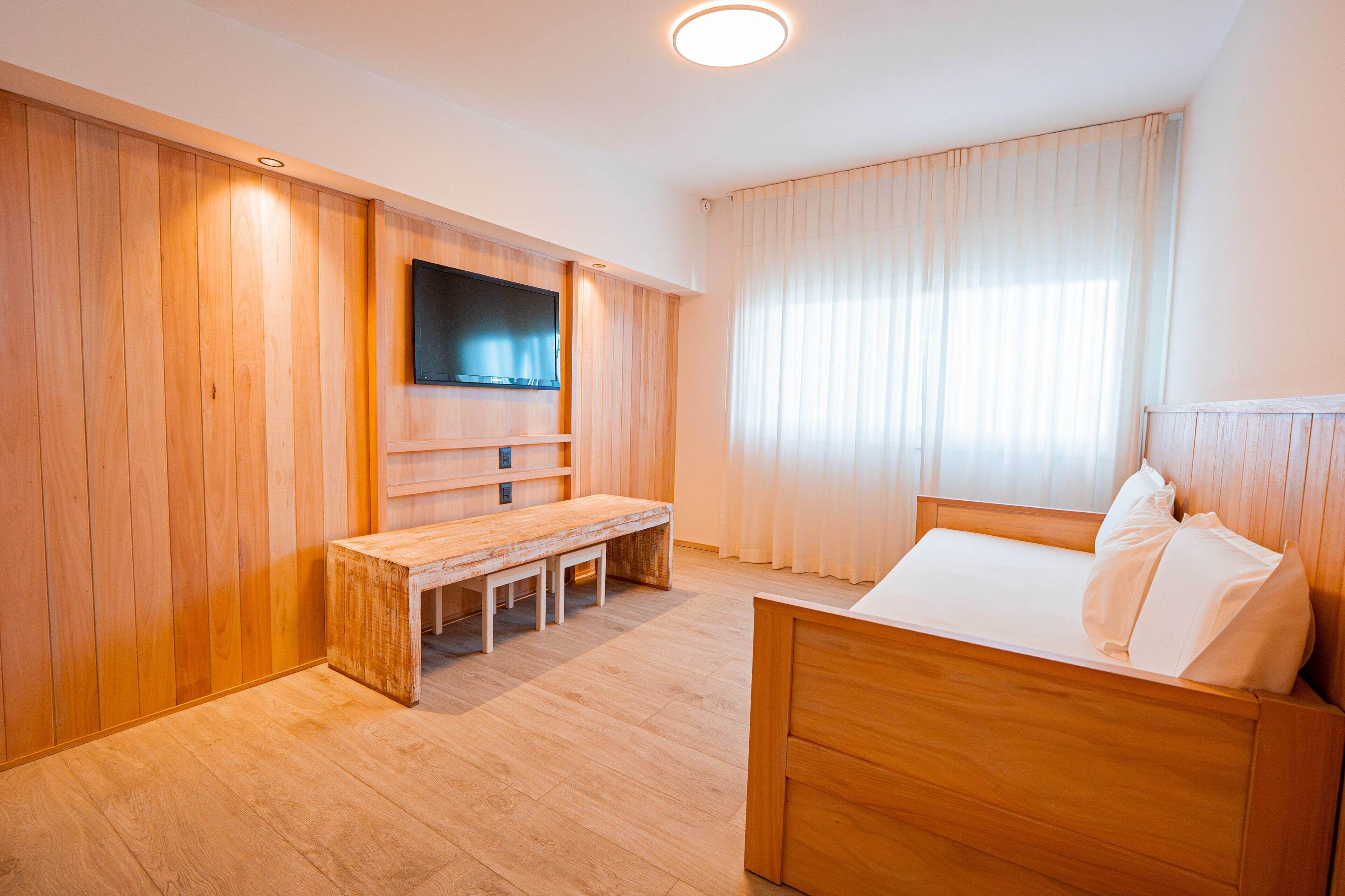 Una habitación de hotel moderna con paneles de madera, un televisor de pantalla plana en la pared, una cama de madera con ropa blanca y un banco con una mesa en el centro.