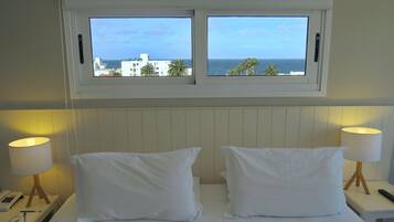 Habitación de hotel con cama, dos lámparas de noche, mesita de noche y ventana con vistas al mar y edificios.