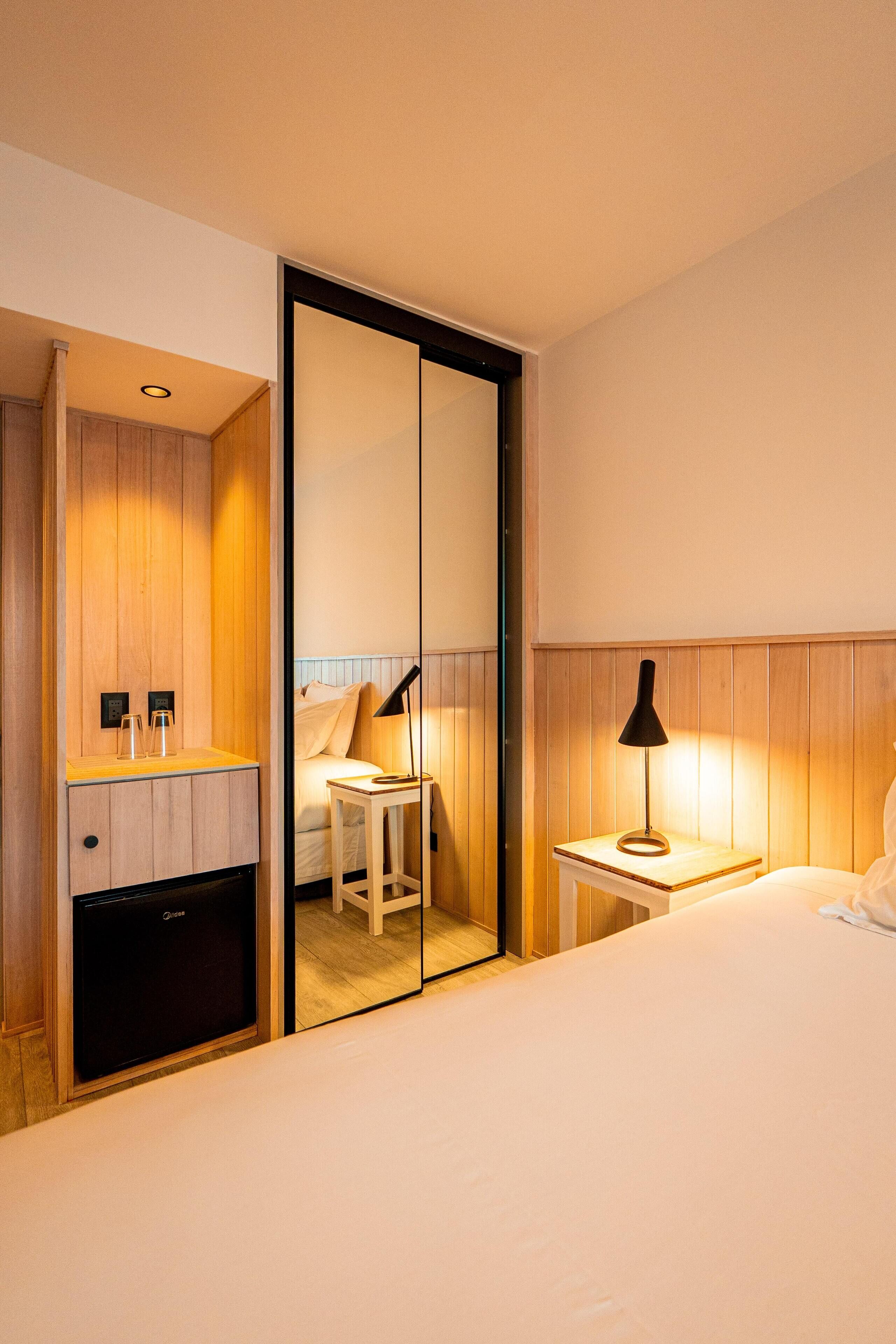 Una habitación de hotel con cama, mesita de noche, lámpara y un espejo que refleja la habitación.