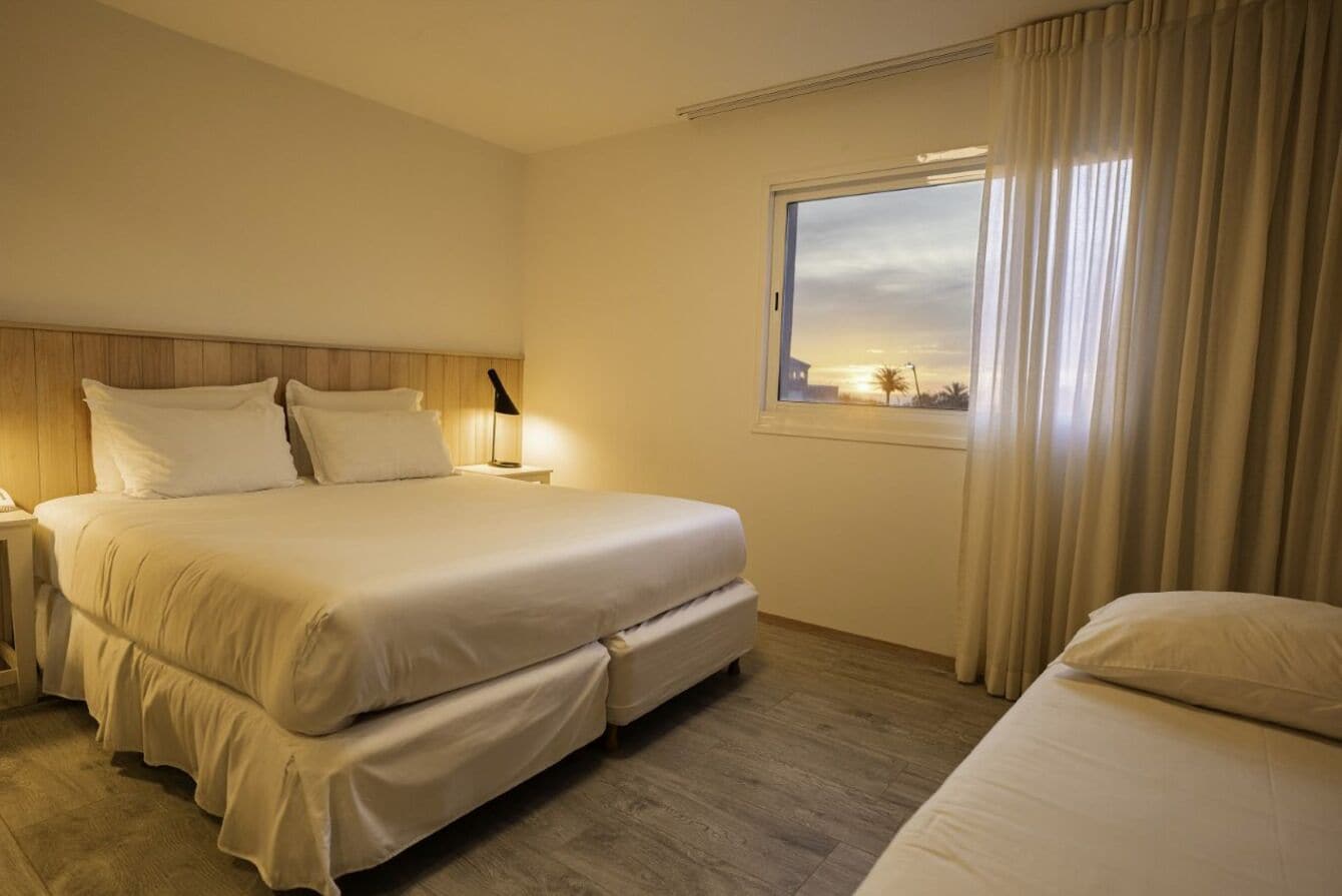 Una habitación de hotel con una cama grande, una mesita de noche con lámpara y una ventana con cortinas.