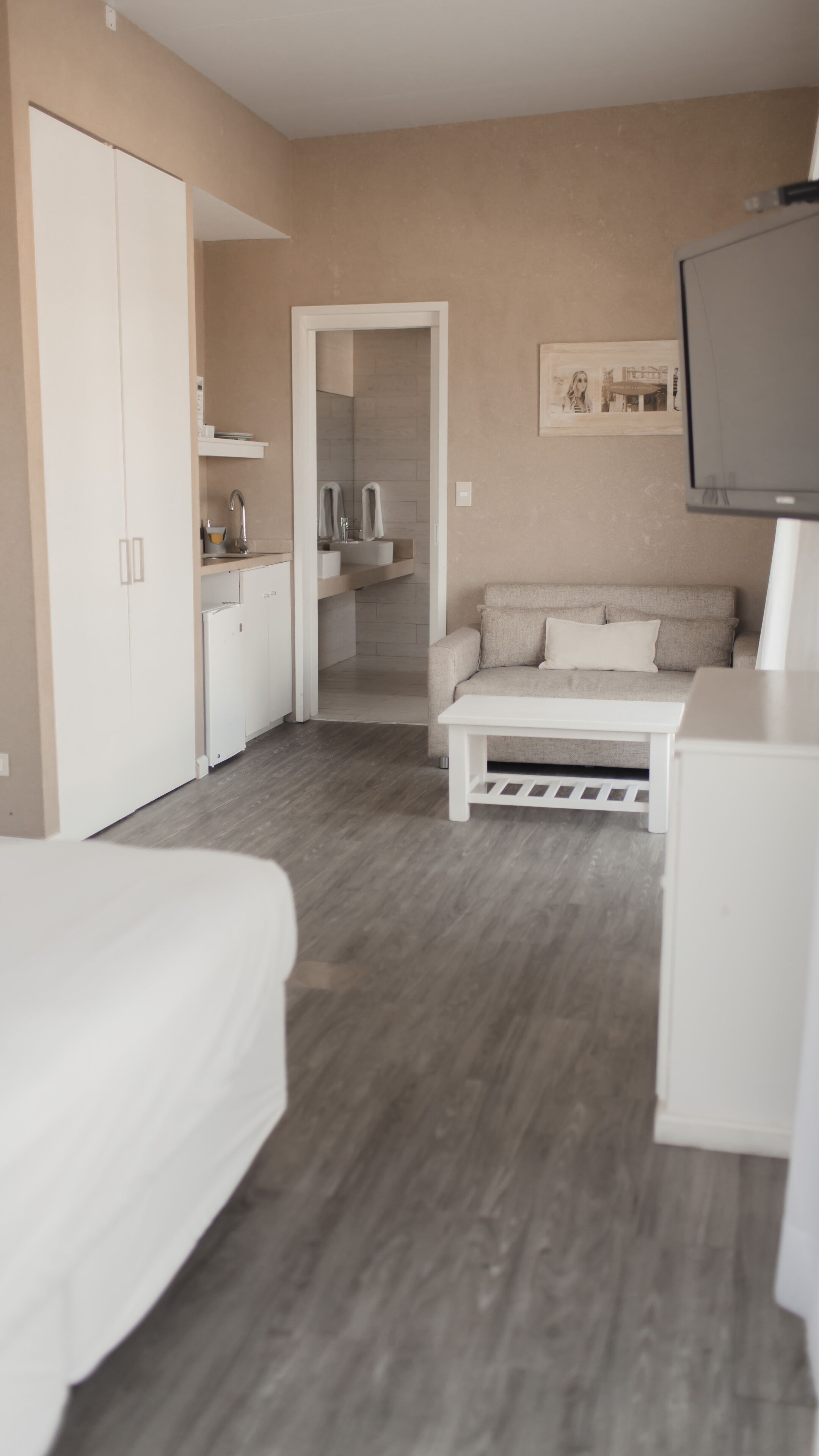 Una habitación de hotel moderna con cama, sofá, televisión, baño con lavamanos y espejo, y un pasillo que conduce a otra habitación.