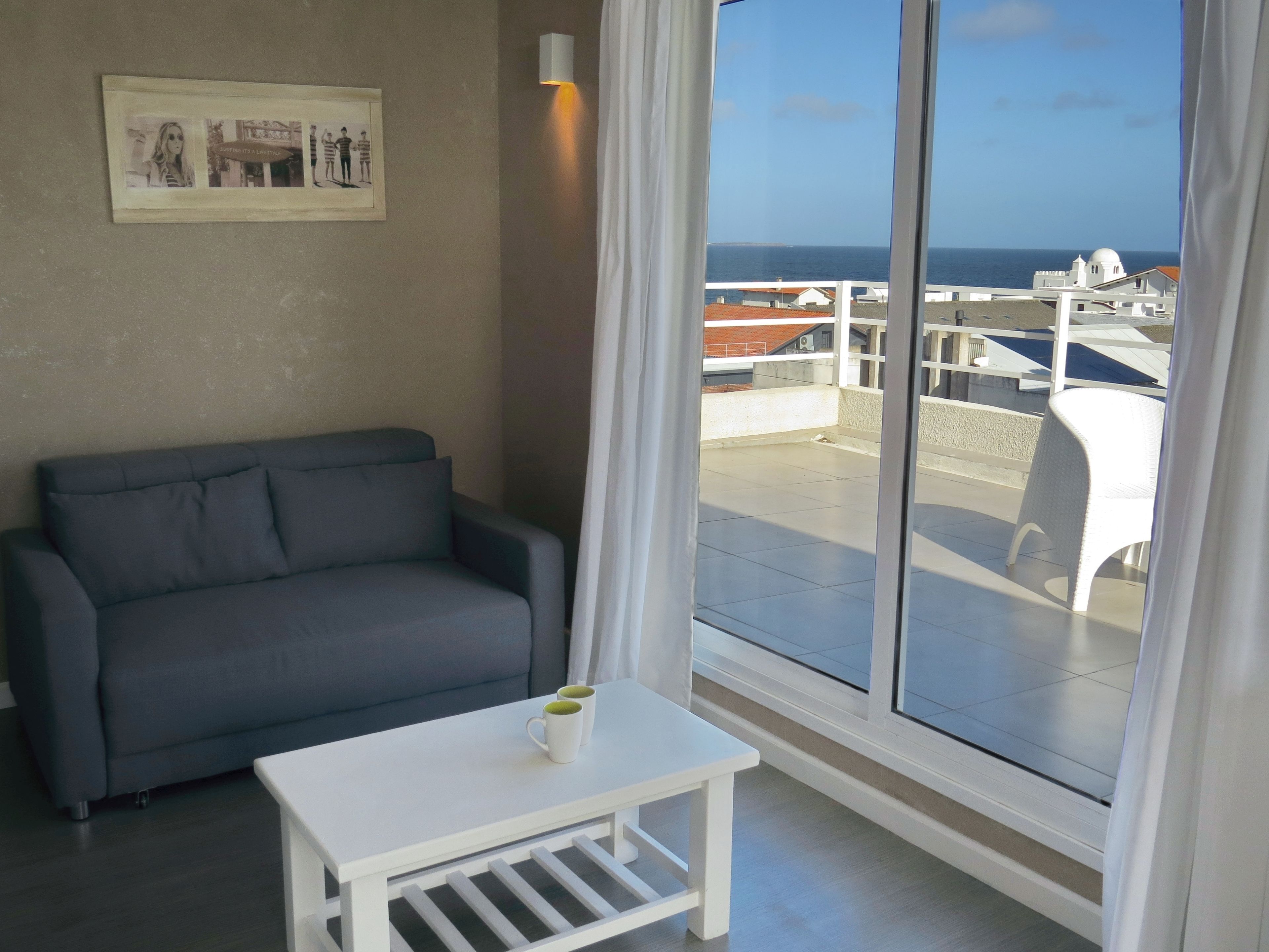 Une pièce avec un canapé gris, une petite table blanche sur laquelle reposent deux tasses, un tableau encadré accroché au mur et une grande fenêtre offrant une vue sur la mer et les bâtiments.