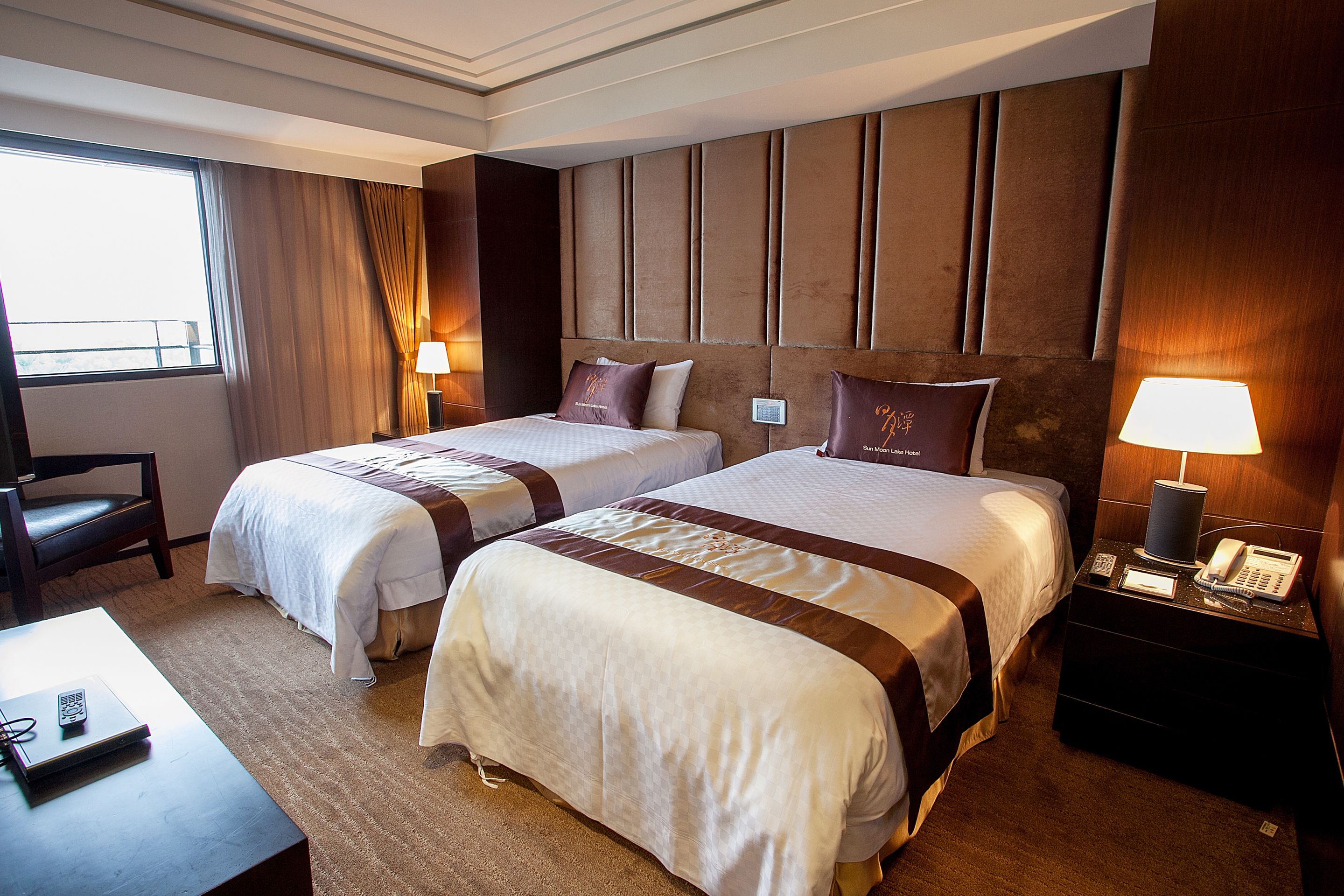 Ett hotellrum med två sängar, ett skrivbord, en stol, ett nattduksbord med en telefon och ett fönster med gardiner.