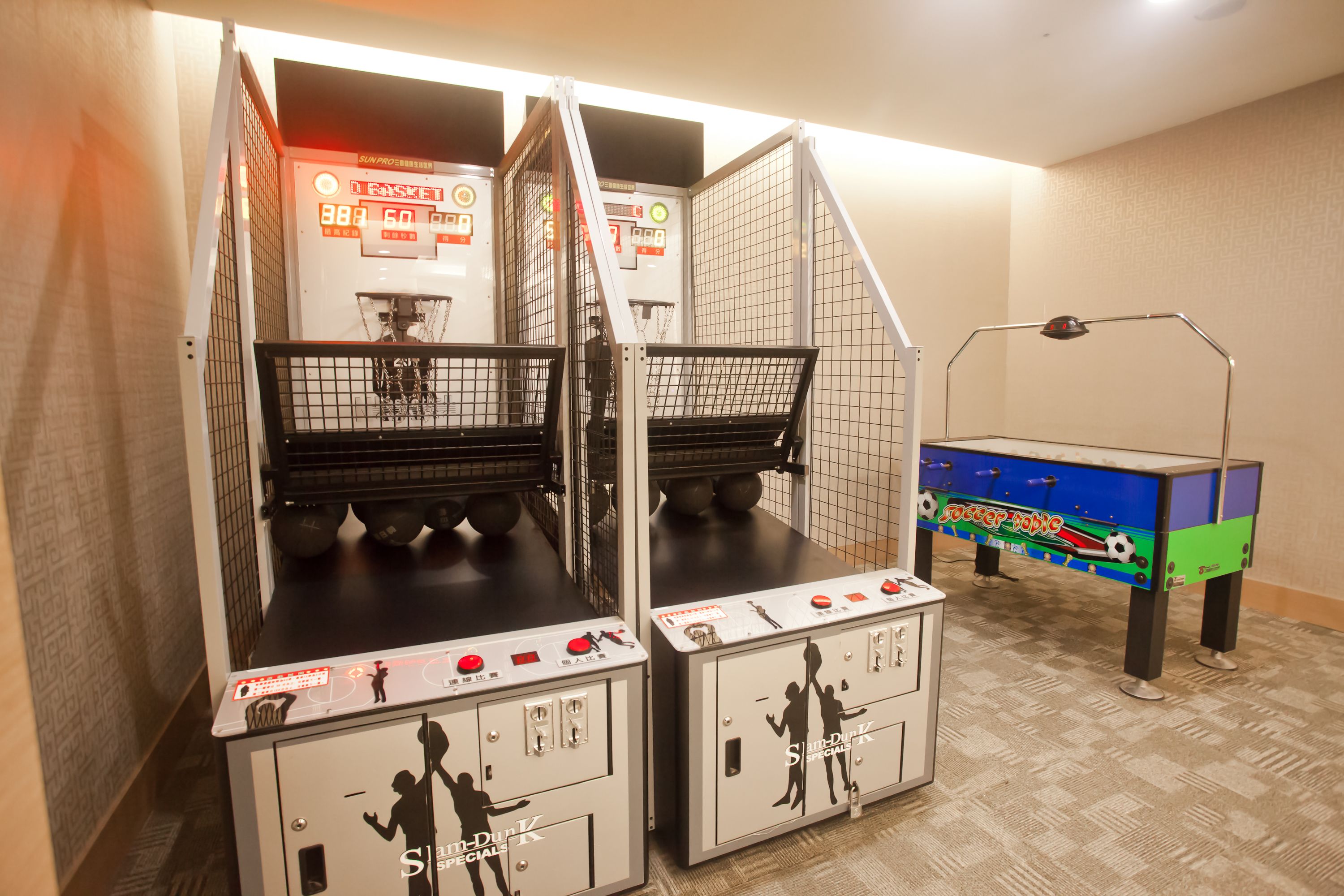 Una habitación con dos mesas de futbolín, una mesa pequeña de ping pong y un aro de baloncesto montado en la pared.