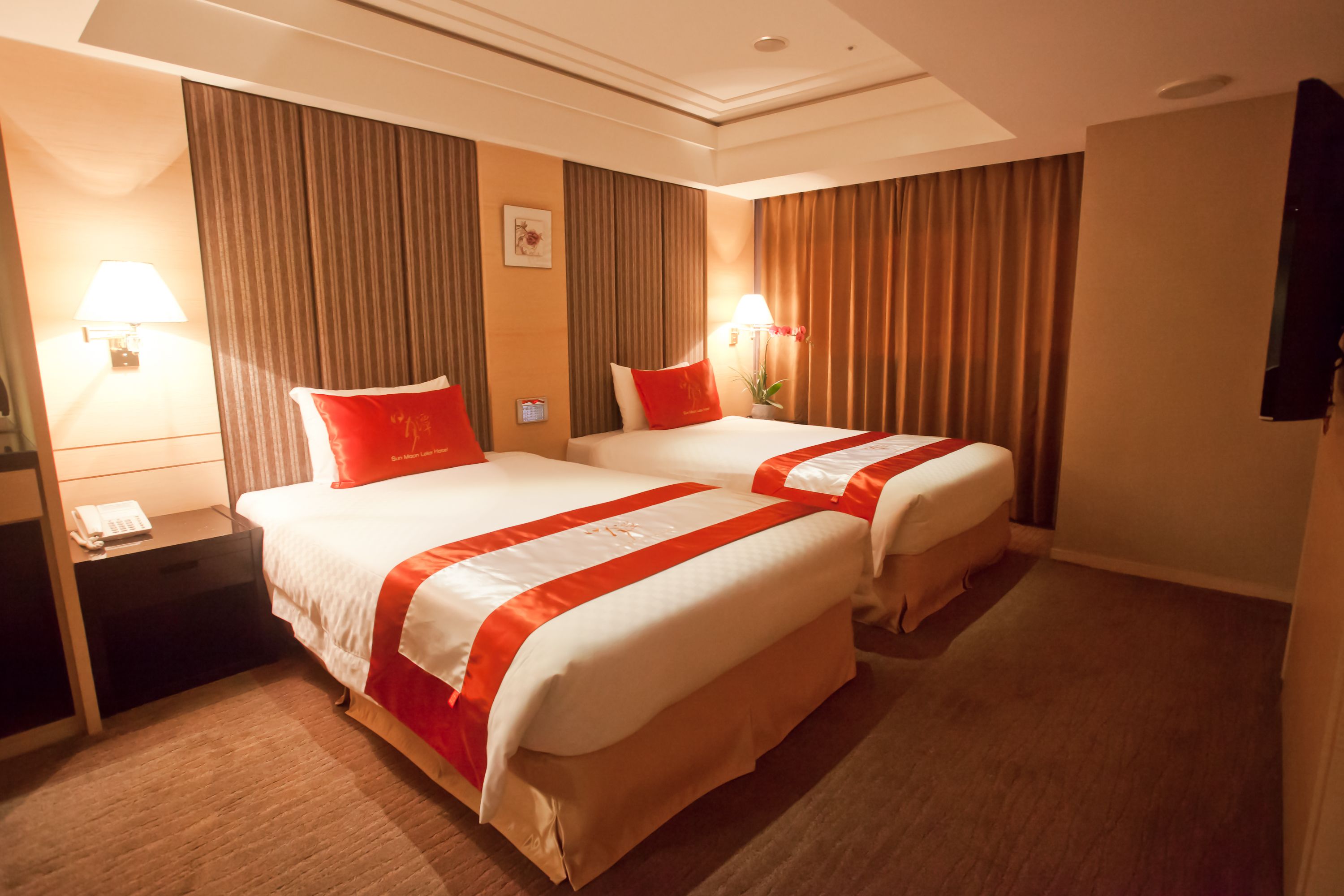 Habitación de hotel con dos camas, cada una con ropa de cama roja y blanca, una mesita de noche con lámpara y un ventanal con cortinas.